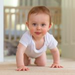 Baby boy in white sleep vest crawling towards the camera on carpet floor.